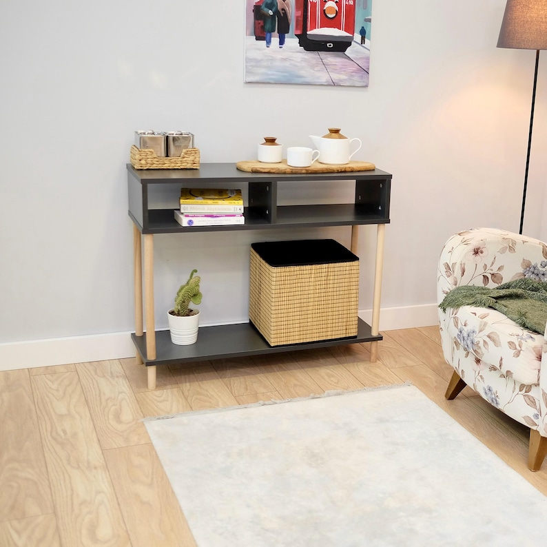 May include: A dark gray console table with two shelves and wooden legs. The table has a woven basket on the top shelf, a small potted plant on the bottom shelf, and a woven storage bin on the lower shelf. The table is in front of a white wall.