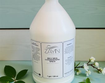 May include: A large white plastic bottle of ZAYN Rice Milk Conditioning Spray. The label indicates the product is 1 gallon / 128 oz. The bottle is set against a light blue wooden background with green leaves and white flowers.
