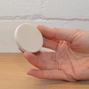 Op de afbeelding: Een klein, rond, gebroken wit object wordt in een hand gehouden. Het object lijkt gemaakt van een vast materiaal, met een glad, licht getextureerd oppervlak. De achtergrond is een lichtgekleurde bakstenen muur en een houten oppervlak.