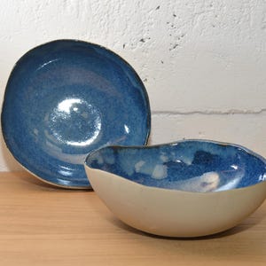 Peut inclure: Deux bols en céramique. L'un est un bol peu profond et ouvert avec un extérieur blanc et un intérieur bleu profond. L'autre est un bol plus grand et rond avec un intérieur bleu similaire. Les deux bols ont une finition brillante.