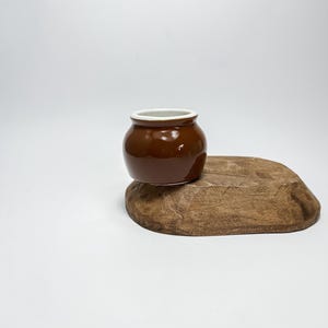 Puede incluir: Una pequeña maceta de cerámica marrón con un borde blanco, sentada sobre un trozo de madera.