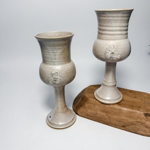 Puede incluir: Dos copas de cerámica hechas a mano con un esmalte beige claro. Cada copa presenta un cuerpo bulboso con un diseño floral y un tallo delgado. Los bordes son acampanados y la superficie tiene un aspecto texturizado. Las copas se exhiben sobre una superficie de madera.