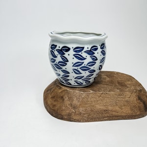 Puede incluir: Un pequeño macetero de cerámica blanca con un patrón de hojas azules y blancas. El macetero tiene un borde festoneado y está sentado sobre una base de madera.