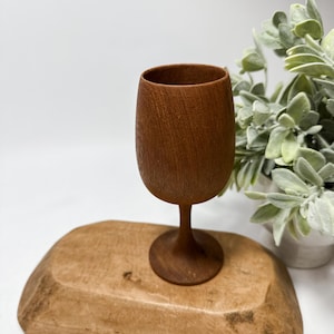 May include: A handcrafted wooden wine glass with a rounded bowl and a slender stem. The glass is made of a warm brown wood and sits on a matching wooden tray. Decorative greenery is in the background.