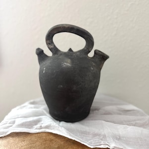 May include: A dark, antique-looking ceramic jug with a rounded body, two spouts, and a large handle. The jug is a dark charcoal color and sits on a white cloth. The jug is likely handmade and has a rustic appearance.