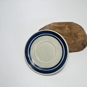 Puede incluir: Un platillo blanco con un diseño de rayas azules y blancas. El platillo está hecho de cerámica y tiene un acabado liso y brillante.