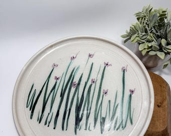 May include: A round, off-white ceramic plate with a hand-painted floral design. The design features green grass-like stems and small purple flowers. The plate has a slightly raised edge and a speckled texture.