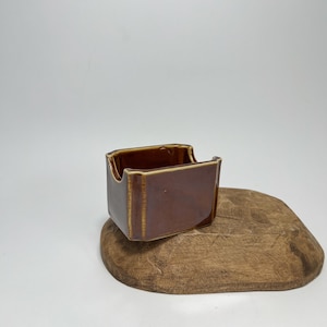 Może przedstawiać: Mały, brązowy ceramiczny dozownik cukru o prostokątnym kształcie. Dozownik ma lekko uniesiony rant wokół górnego otworu.