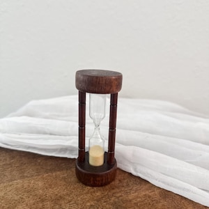 May include: A small hourglass with a dark brown wooden frame and base. The hourglass contains light-colored sand and clear glass. The hourglass is approximately 4 inches tall and sits on a wooden surface.