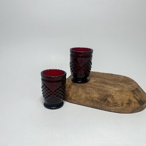 Op de afbeelding: Twee kleine, robijnrode glazen vaasjes met een diamantpatroon. De vaasjes staan op een houten oppervlak.