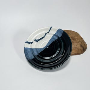 Puede incluir: Un plato de cerámica blanco y negro con un esmalte azul. El plato tiene un diseño ondulado y está ligeramente inclinado.