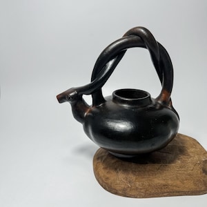 May include: A black ceramic teapot with a unique twisted handle. The teapot has a rounded body and a small spout. It is sitting on a wooden base.