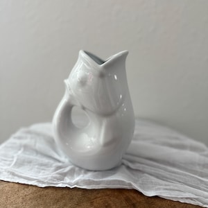 May include: A white ceramic jug with an unusual, organic form. The jug features a curved handle and a flared spout. The interior of the jug is a darker grey colour. The jug rests on a white cloth.