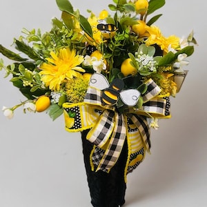 May include: A decorative floral arrangement featuring yellow sunflowers, lemons, and greenery. The arrangement is accented with a black and white checkered bow and a bee-shaped embellishment. The vase is black.