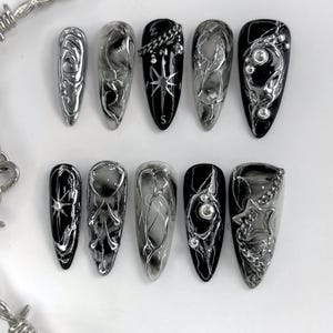 May include: A set of ten long, almond-shaped artificial nails. The nails feature a mix of silver metallic and black designs, including chains, swirls, and starbursts. Some nails have a smoky, marbled effect, while others are solid black.