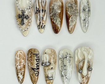 May include: A set of ten almond-shaped, press-on nails with various designs. The nails feature a mix of cream, brown, and silver colors. Some nails have cross, angel, and candle designs, while others have text that reads "born to die" and "angel".