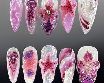 May include: Ten pink and purple nail tips with different sea-themed designs. The designs include a jellyfish, a starfish, a seashell, and a flower.