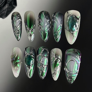 Puede incluir: Diez uñas postizas largas y ovaladas con un acabado verde e iridiscente. Cada uña tiene un diseño diferente con remolinos y líneas plateadas.