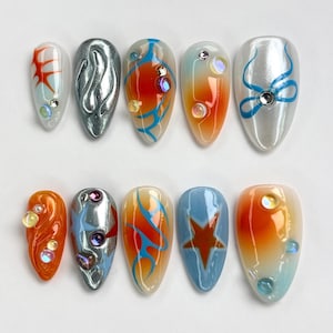 May include: Set of ten false nails with various designs. The nails are almond shaped and feature a mix of orange, blue, silver, and white colors. Some nails have a gradient effect, while others have abstract designs, stars, and swirls.