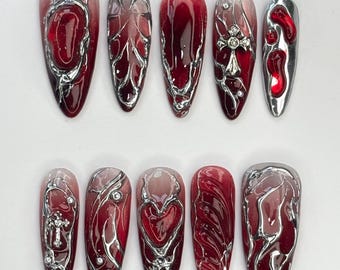 Puede incluir: Un juego de diez uñas postizas largas en forma de almendra. Cada uña presenta un degradado de rojo y plata, con intrincados diseños y adornos plateados, incluyendo cruces y corazones. Las uñas tienen un acabado brillante.