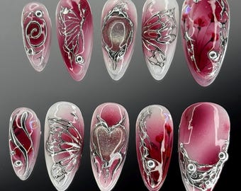 Puede incluir: Diez uñas postizas largas en forma de almendra con un degradado de blanco a rojo. Cada uña tiene un diseño plateado diferente, incluyendo mariposas, corazones y espirales.