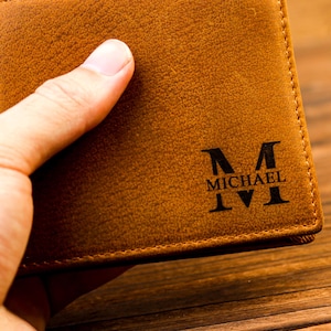 Puede incluir: Una cartera de cuero marr&oacute;n con una inicial "M" grabada y el nombre "MICHAEL" en la parte delantera.