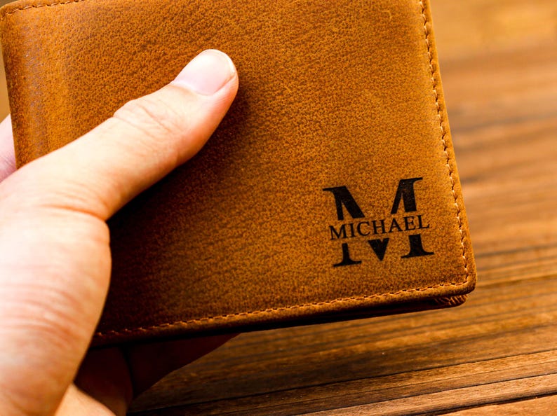 Puede incluir: Una cartera de cuero marr&oacute;n con una inicial "M" grabada y el nombre "MICHAEL" en la parte delantera.