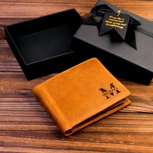 Puede incluir: Una cartera de cuero marr&oacute;n con un monograma personalizado. El monograma es en negro y dice "MICHAEL M". La cartera est&aacute; en una caja de regalo negra con una cinta negra y una etiqueta dorada.