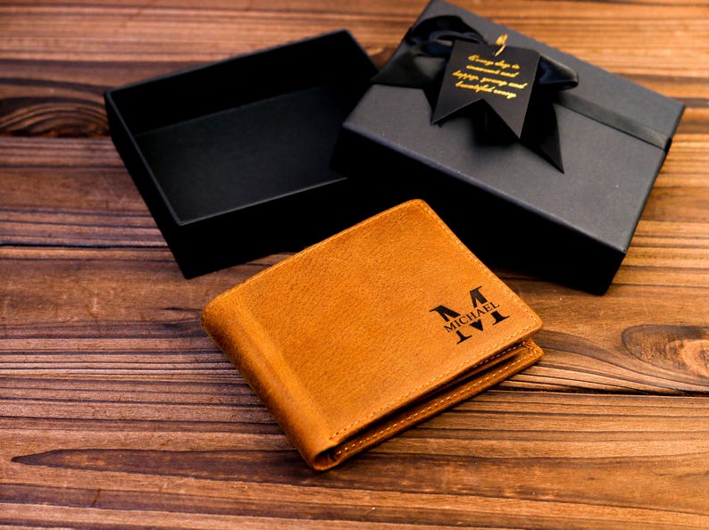 Puede incluir: Una cartera de cuero marr&oacute;n con un monograma personalizado. El monograma es en negro y dice "MICHAEL M". La cartera est&aacute; en una caja de regalo negra con una cinta negra y una etiqueta dorada.