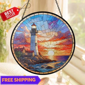 May include: Round stained glass window art depicting a lighthouse at sunset. The lighthouse is white with a red roof, set against a colorful sky of blues, oranges, and purples. The image is framed in black and hangs from a chain.