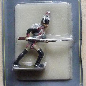 Könnte beinhalten: Eine verpackte Spielzeug-Soldatenfigur aus Plastik, in Rot, Weiß und Schwarz bemalt und mit einem Gewehr bewaffnet. Die Verpackung trägt den Text "Authentique Soldat de Plomb Modele de Collection".