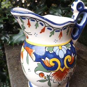 Op de afbeelding: Een witte keramische kan met een blauw, geel, oranje en groen bloemenmotief. De kan heeft een handvat met een decoratieve lus.