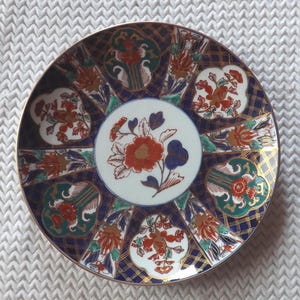 Può includere: Un piatto rotondo in ceramica con un medaglione centrale bianco decorato da un motivo floreale rosso e blu. Il bordo è diviso in pannelli alternati con motivi a griglia blu scuro e sezioni bianche decorate con fiori colorati.