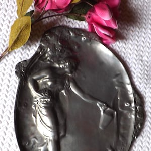 Può includere: Un vassoio ovale in metallo color argento con un design in rilievo raffigurante una figura che tiene un secchio, accompagnato da un ramo di fiori artificiali rosa con foglie verdi. Il vassoio ha una superficie liscia e riflettente con dettagli in rilievo.