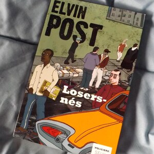 Peut inclure: Couverture du livre "Elvin Post Losers-nés" avec une illustration de personnes dans une scène de rue avec une voiture orange. Le titre du livre est en grandes lettres noires en gras. Le livre est sur une surface bleue.