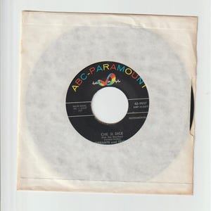 Puede incluir: Un disco de vinilo de 45 RPM vintage en una funda de papel. La etiqueta del disco es negra con texto blanco y el logotipo de ABC-Paramount en un diseño de arcoíris. El título "CHE SI DICE" es visible.