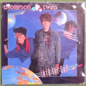 May include: A vintage record album cover featuring a band called "Thompson Twins". The album cover is blue and features three people, two men and one woman. The album title is "Into The Gap".
