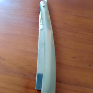 Op de afbeelding: Een vintage recht scheermes met een crèmekleurige handgreep en een zilveren lemmet. Het lemmet is gedeeltelijk open en onthult de scherpe rand. De handgreep heeft een afgerond uiteinde en is vastgezet met metalen klinknagels. De tekst "T.R. CADMAN SHEFFIELD" is zichtbaar op het lemmet.