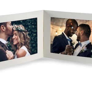 May include: A white, bi-fold photo frame displaying two wedding photographs. One shows a couple, the other a same-sex couple. A silver ribbon is attached to the frame.