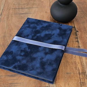 May include: A navy blue velvet journal with a matching ribbon closure. The journal is rectangular and has a soft, textured appearance. A dark, round decorative object is in the background. The journal is on a wooden surface.