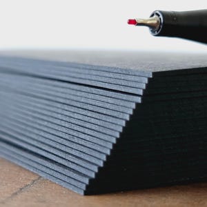 Puede incluir: Una pila de tarjetas de papel negro con un bolígrafo en la esquina superior derecha. El bolígrafo tiene un cuerpo negro y un agarre amarillo. La punta del bolígrafo es roja. Las tarjetas de papel están apiladas en diagonal.