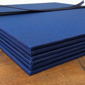 May include: A stack of six navy blue fabric-covered books, bound together with a black ribbon. The books are rectangular and have a textured surface. The image is taken on a wooden surface.