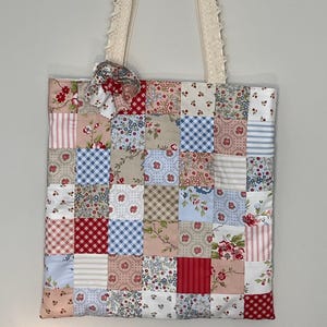 Könnte beinhalten: Eine Patchwork-Tragetasche mit einem cremefarbenen, mit Spitze besetzten Griff. Der Taschenkorpus ist ein Quilt aus verschiedenen floralen und gemusterten Stoffen in Rot, Rosa, Blau und Creme. Eine kleine Stoffschleife ziert die Oberseite.