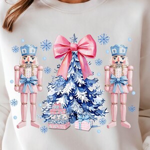 May include: White sweatshirt with a watercolor design of a Christmas tree with a pink bow, two pink nutcracker figurines, and blue and white gift boxes.