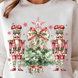 May include: White sweatshirt with a Christmas tree and two nutcracker soldiers printed on the front. The nutcrackers are wearing red and gold uniforms and holding a candy cane and a drum. The tree is decorated with a red bow and a star.
