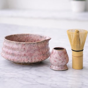 Op de afbeelding: Een matcha-theeset met een roze en witte keramische kom met een tuit, een kleine bijpassende keramische beker en een bamboe klopper. De set is ontworpen voor het bereiden van matcha-thee.