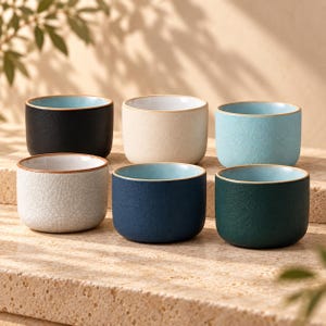 Könnte beinhalten: Ein Set aus sechs kleinen Keramikbechern mit abgerundeten Kanten. Die Becher haben verschiedene Außenfarben, darunter Schwarz, Creme, Hellblau, Dunkelblau, Dunkelgrün und eine weiße Craquelé-Glasur. Jeder Becher hat eine glatte, hellblaue Innenseite.