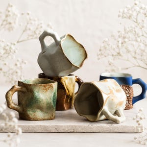 Puede incluir: Una colección de tazas de cerámica hechas a mano en varios colores y texturas. Las tazas presentan formas irregulares únicas, con colores como verde, marrón, azul y crema. Algunas tazas tienen exteriores texturizados y colores interiores contrastantes.
