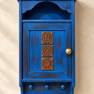 Könnte beinhalten: Ein wandmontierter blauer Holzschrank mit abgenutzter Oberfläche, ausgestattet mit einem offenen oberen Regal, einer zentralen Tür mit drei quadratischen, rostfarbenen Zierfliesen aus Metall, einem runden Messingknopf und drei Haken.