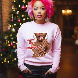 Peut inclure: Un sweat-shirt rose clair avec un graphisme d'une femme tenant un bonbon à la menthe poivrée. Le graphisme comprend les mots "Pretty Paid & Peppermint" en rouge. La femme porte des créoles dorées et un collier.